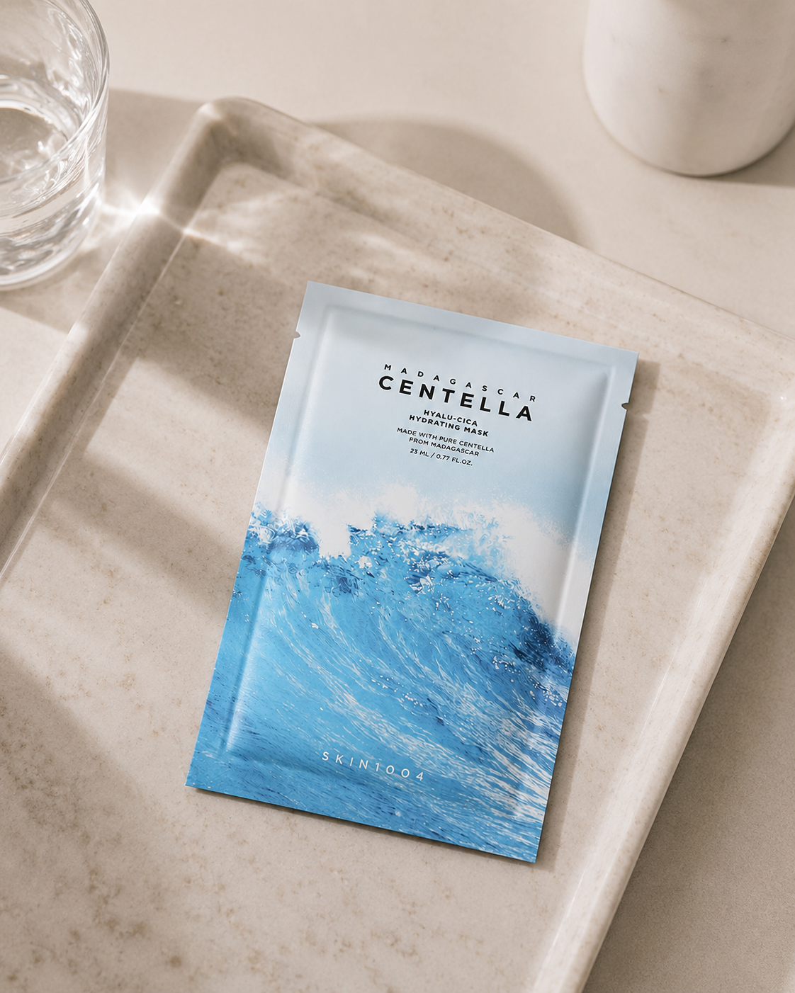 Skincare sheet mask on a beige tray with a glass of water and white cup in the background