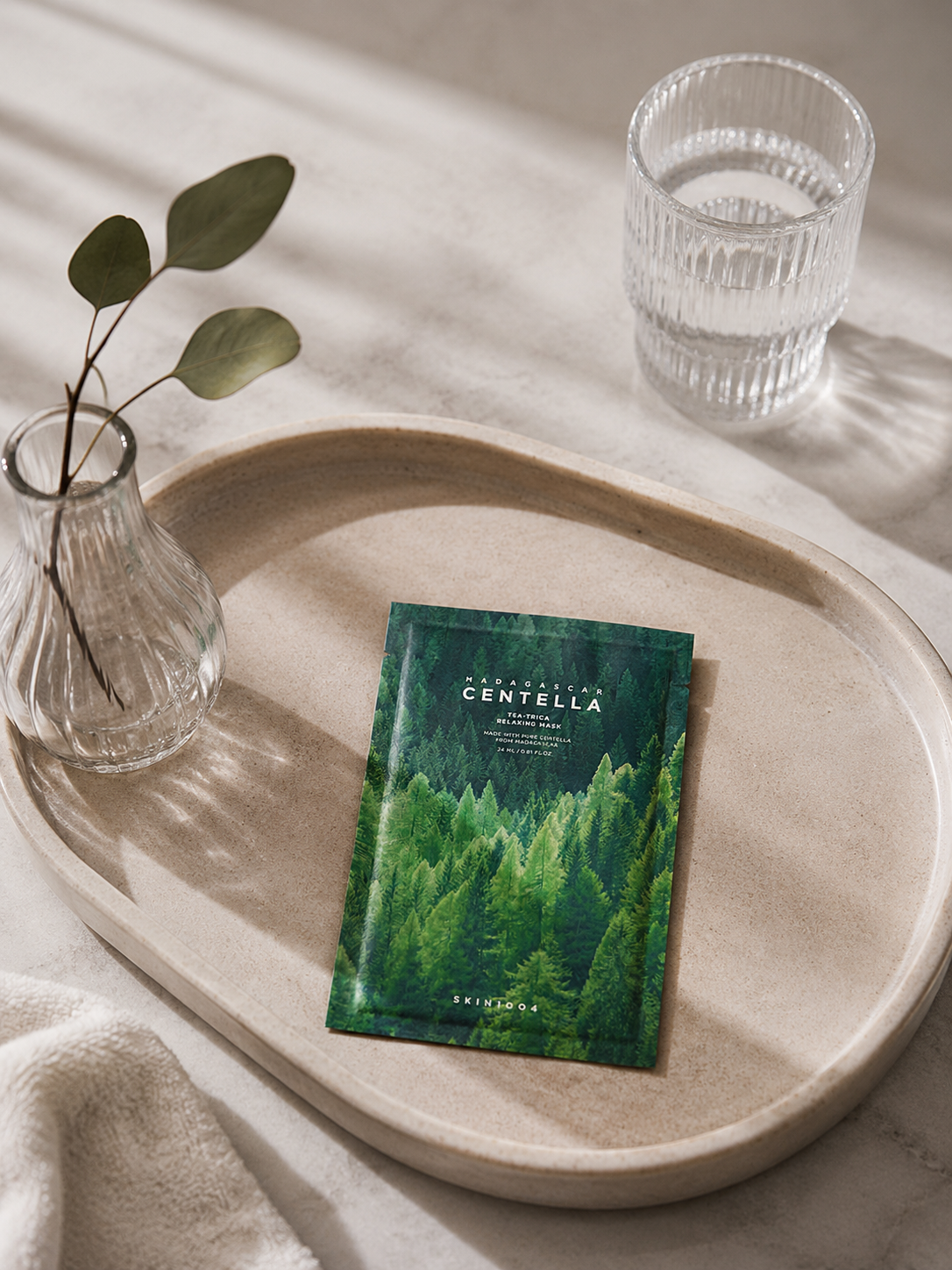 Green skincare sheet mask on a wooden tray with a glass of water and a vase with leaves.