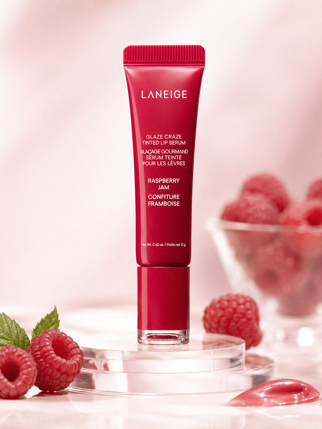A tube of Laneige Glaze Craze Tinted Lip Serum in Raspberry Jam stands upright, surrounded by fresh raspberries and green leaves, emphasizing its vibrant red packaging and glossy finish.